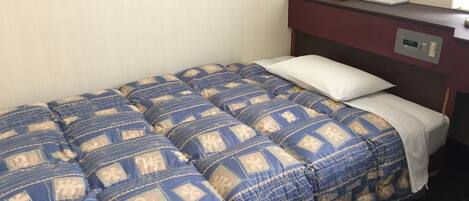 Uma cama de solteiro com um edredom azul e bege estampado, um travesseiro branco e uma mesinha de cabeceira com um estojo de lenços.