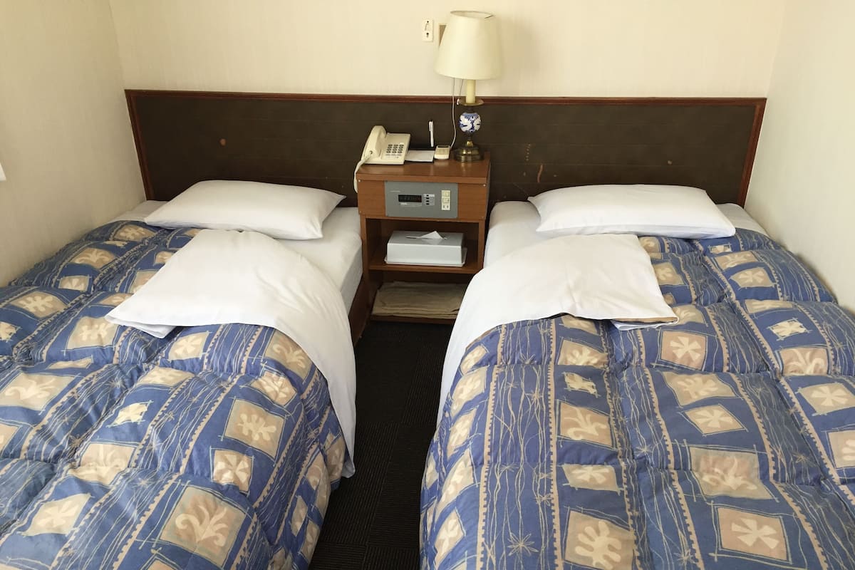 A hotel room with two beds, each with patterned bedding, a bedside table with a lamp, and a digital clock.
