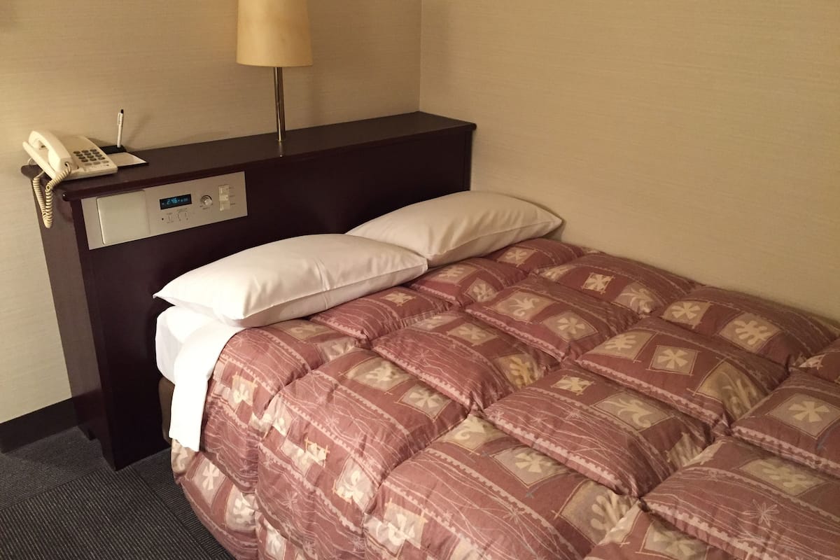 A hotel room with a bed, a bedside table with a lamp, a phone, and a digital clock.