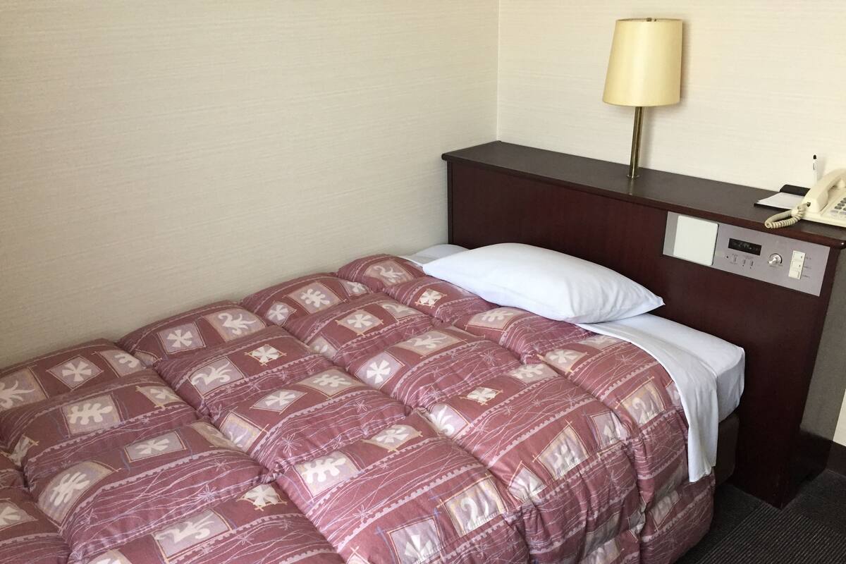 A hotel room with a neatly made bed, a bedside table with a lamp, a phone, and a control panel.