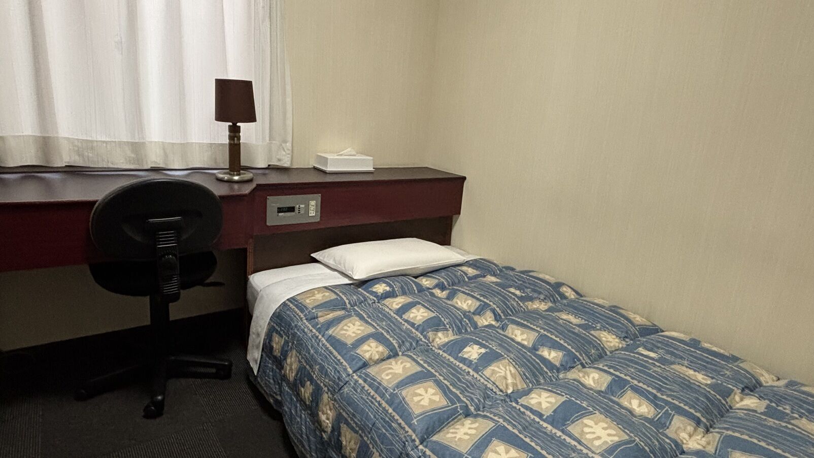 Une chambre d’hôtel avec un lit, un bureau, une chaise, une lampe et une fenêtre avec des rideaux.