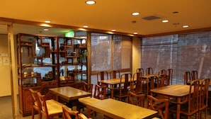 A restaurant with wooden tables and chairs, a decorative wooden shelf with various items, and large windows with blinds. - Kochi Business Hotel Bekkan (Kochi)