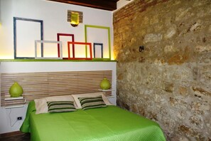 Un dormitorio con cama, almohadas, una mesita de noche, un estante con marcos y una pared de piedra.