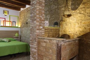 Una habitación rústica con paredes de ladrillo visto, techo de madera, cama con ropa de cama verde, lámpara colgante y un pequeño estante con objetos decorativos.