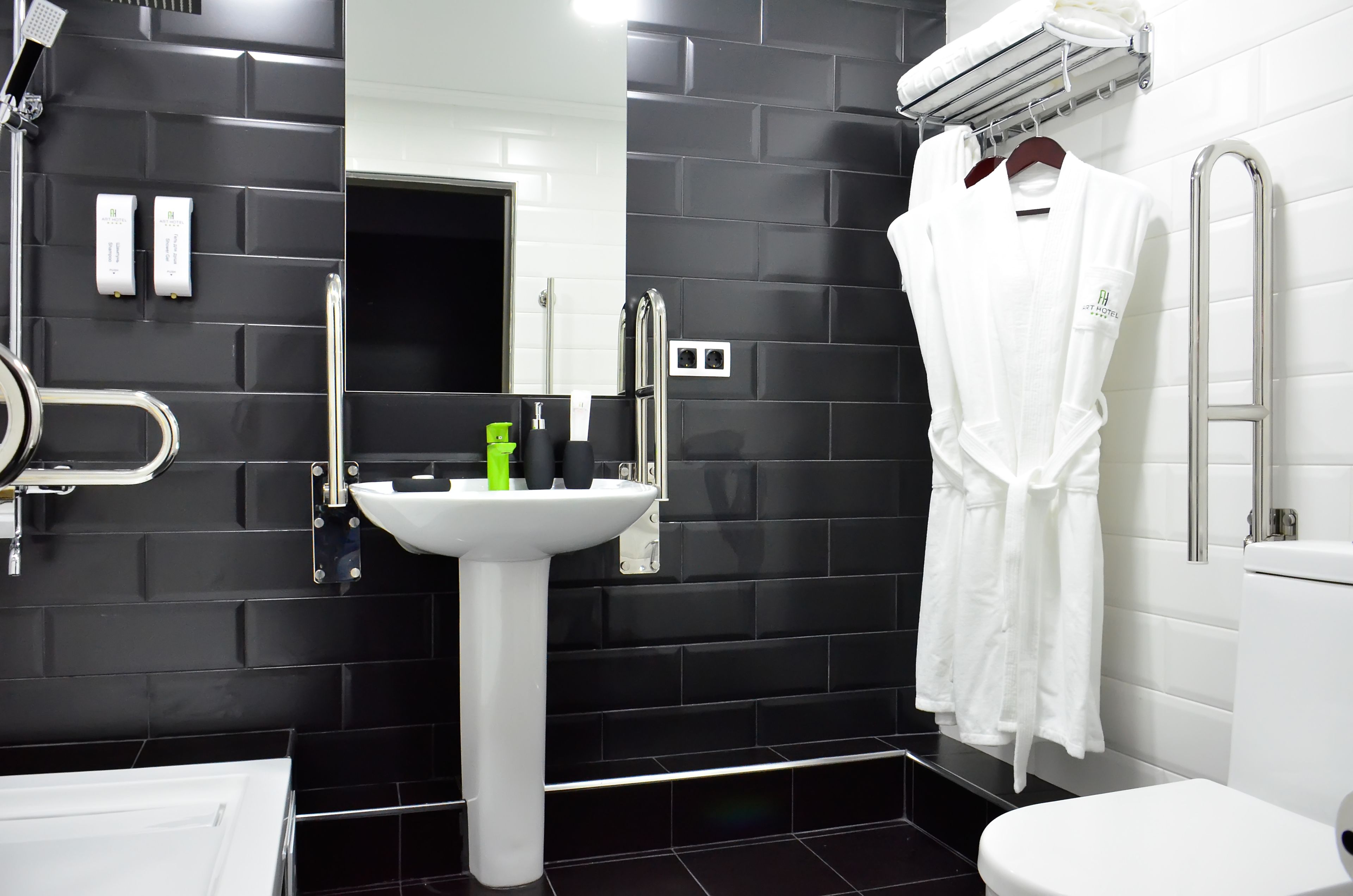 Et moderne badeværelse med vægge af sorte fliser, en hvid vask og et hvidt toilet. Der hænger en hvid badekåbe på et stativ, og der er en grøn flaske på vasken.