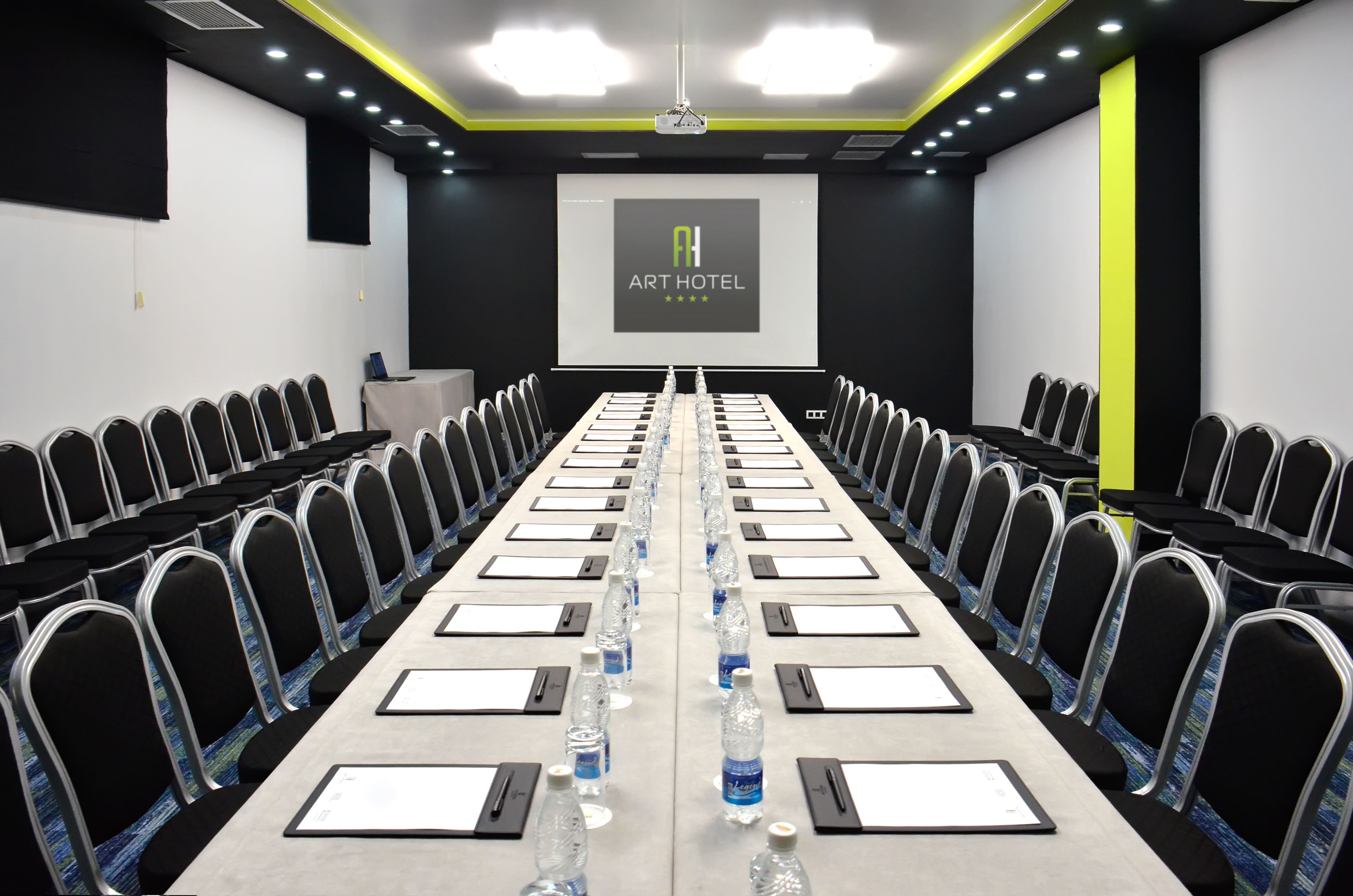 A conference room with a long table, chairs, water bottles, and a projector screen displaying "ART HOTEL".