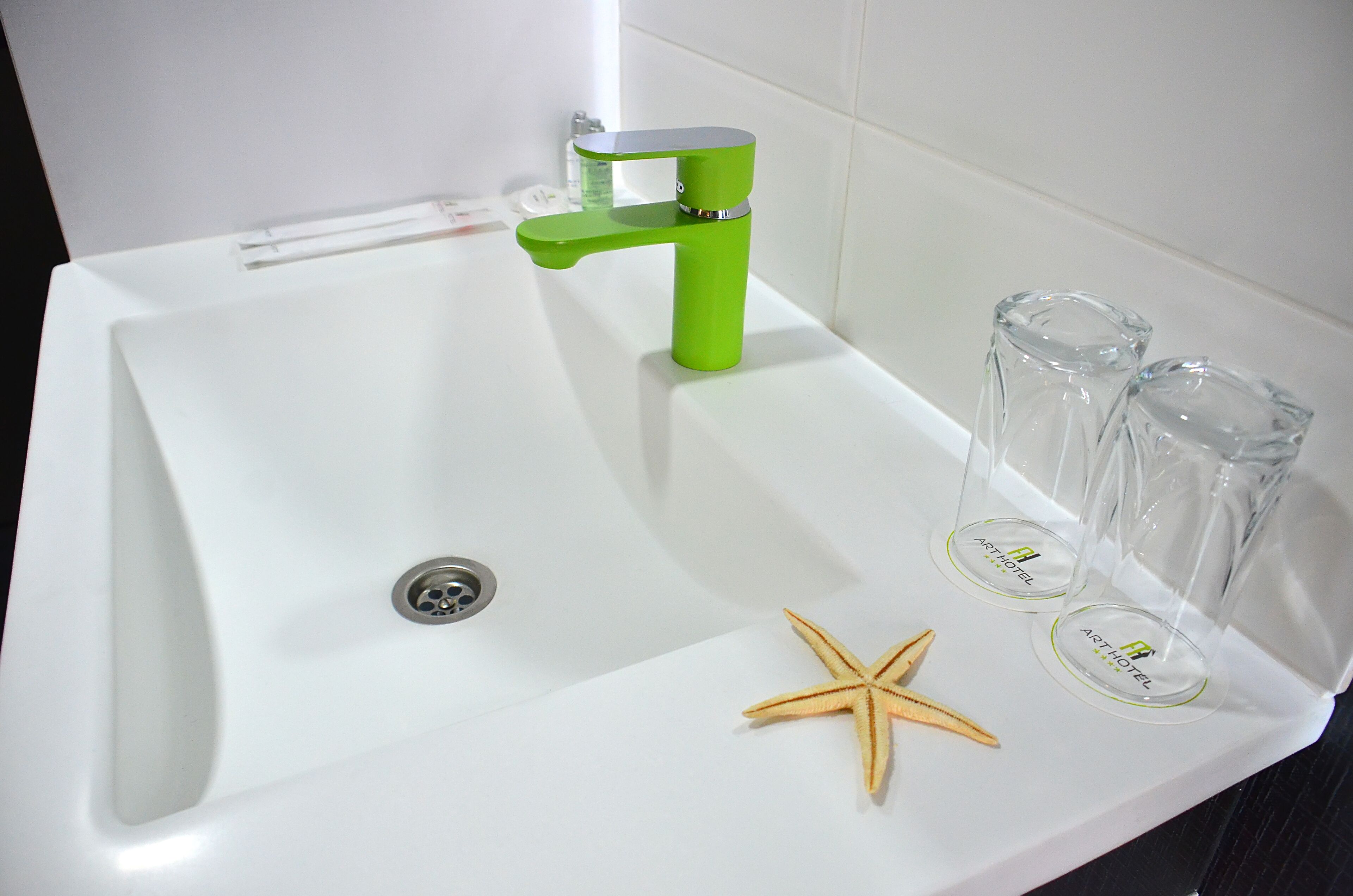 A white bathroom sink with a green faucet, a starfish decoration, and three glasses with the hotel name.