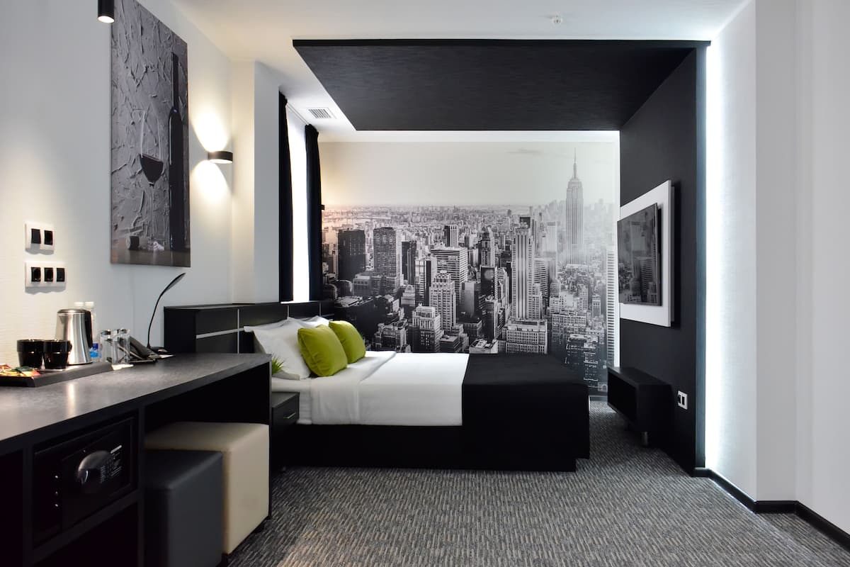 Een moderne hotelkamer met een stadsmuur op de muur, een bed met wit beddengoed en groene kussens, een bureau met een koffiezetapparaat, en een klein zithoekje.