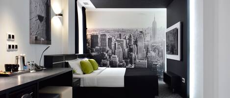 Une chambre d’hôtel moderne avec une fresque représentant un paysage urbain sur le mur, un lit avec une literie blanche et des oreillers verts, un bureau avec une machine à café et un petit coin salon.
