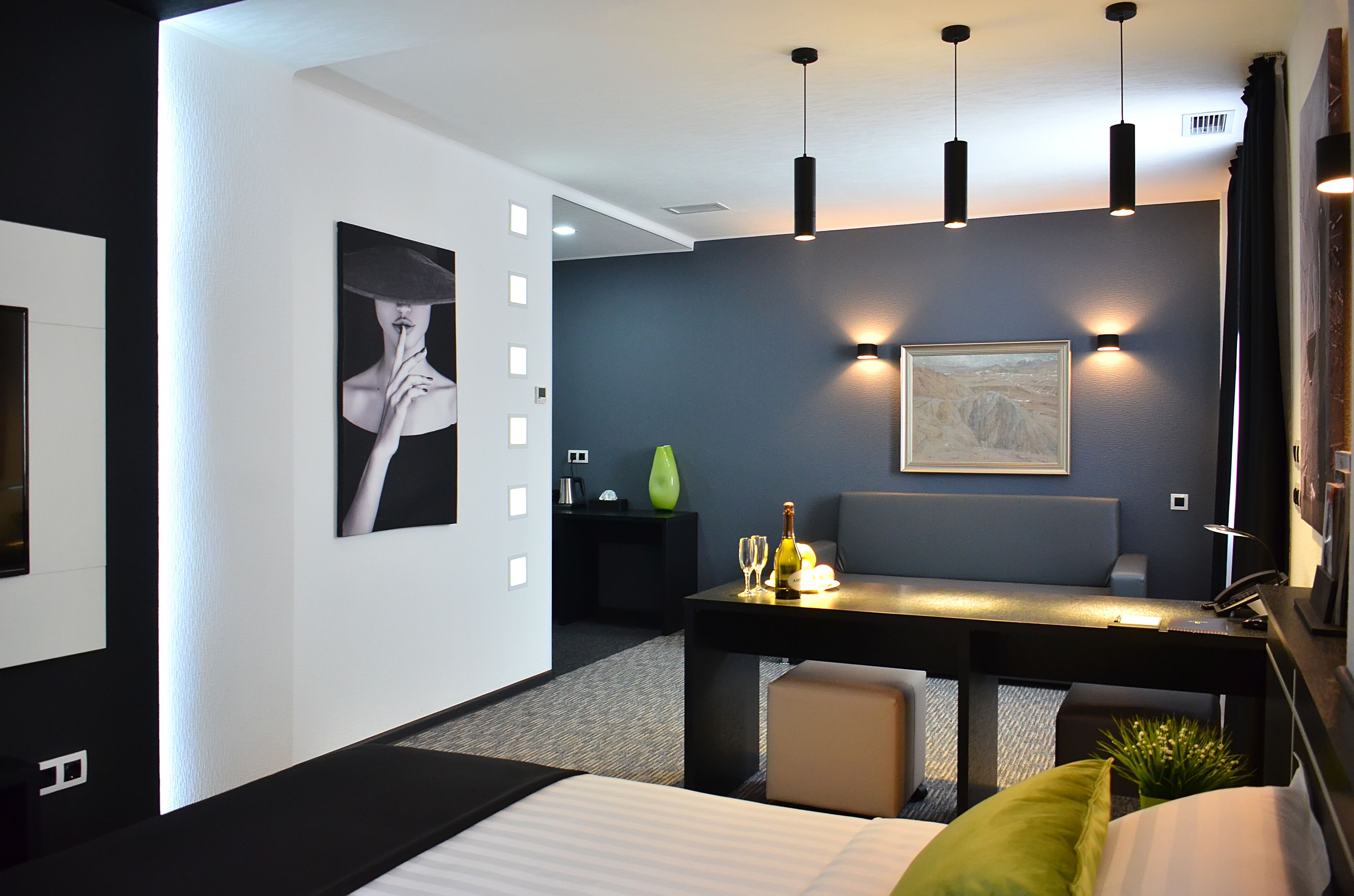A modern hotel room with a bed, a desk with a chair, a small table with a bottle and glasses, and a painting on the wall.