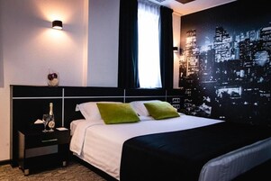 Habitación de hotel con una cama, una mesita de noche con una botella y vasos, una lámpara de pared y un mural de un paisaje urbano.