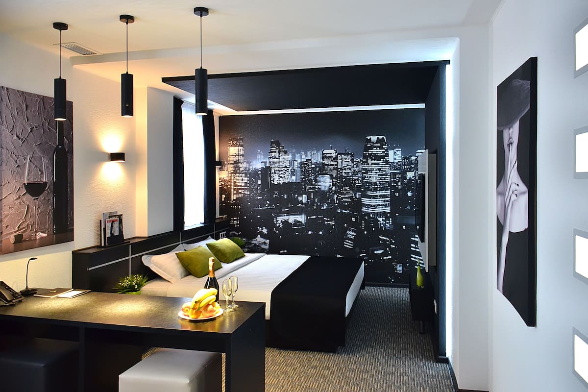 Een moderne hotelkamer met een stadslandschap op de muur, een bureau met een laptop en een bed met kussens.