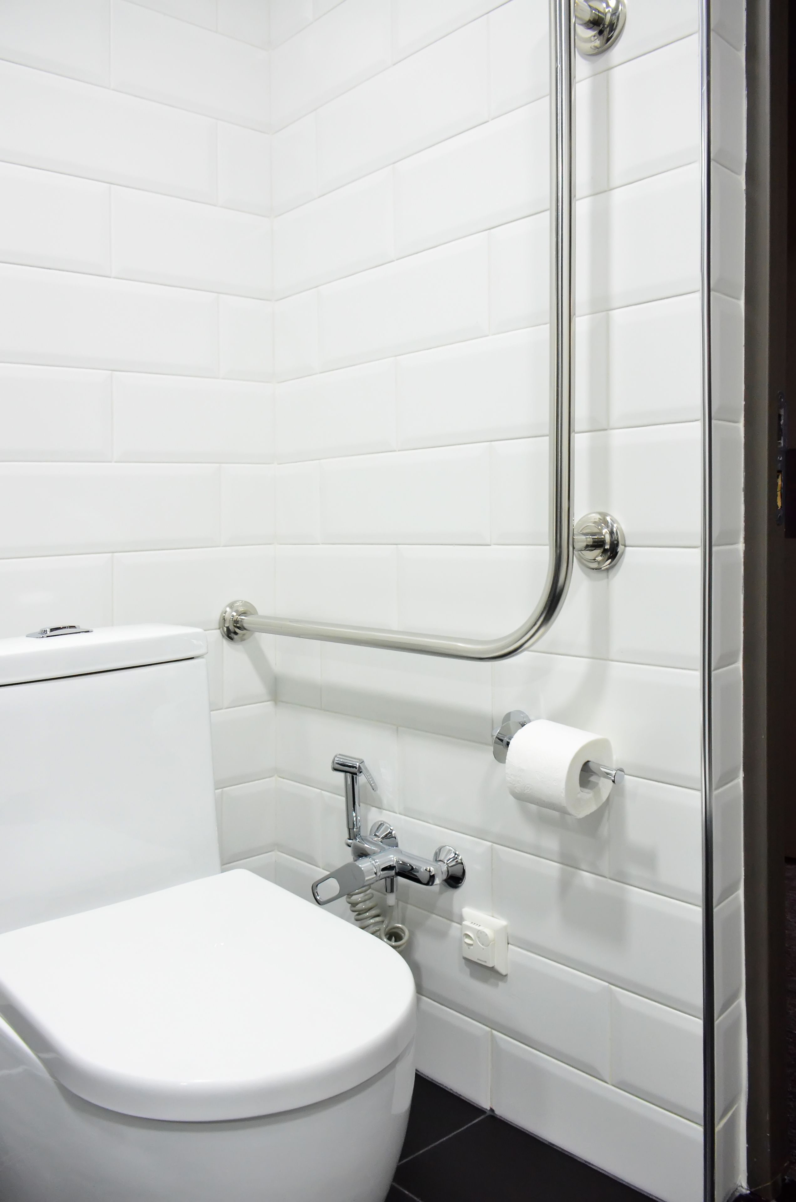 Une salle de bain moderne avec des carreaux blancs, des toilettes blanches, une barre d’appui et un rouleau de papier toilette fixé au mur.
