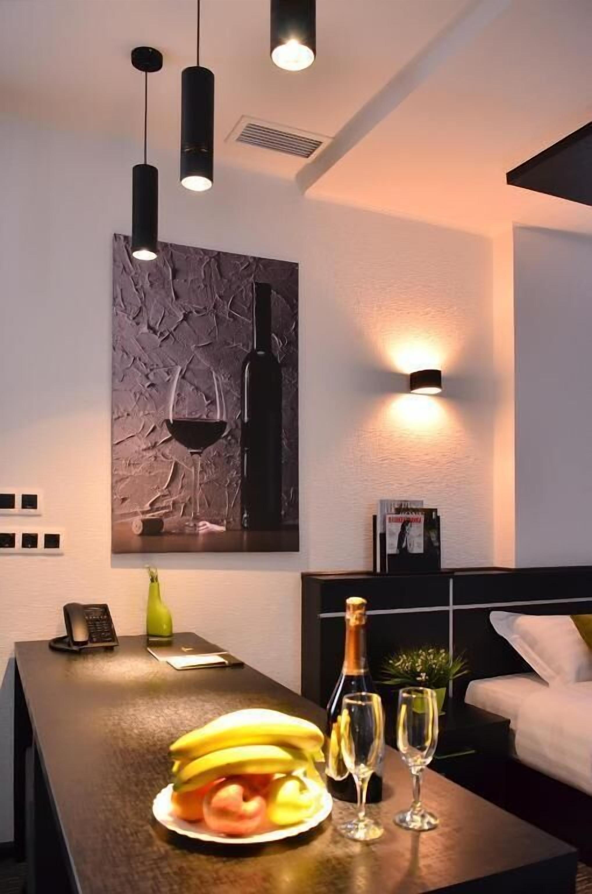 Habitación de hotel moderna con un escritorio de madera oscura, una botella de vino, dos copas y un plato de plátanos. Hay un cuadro en la pared y una planta pequeña en la mesita de noche.