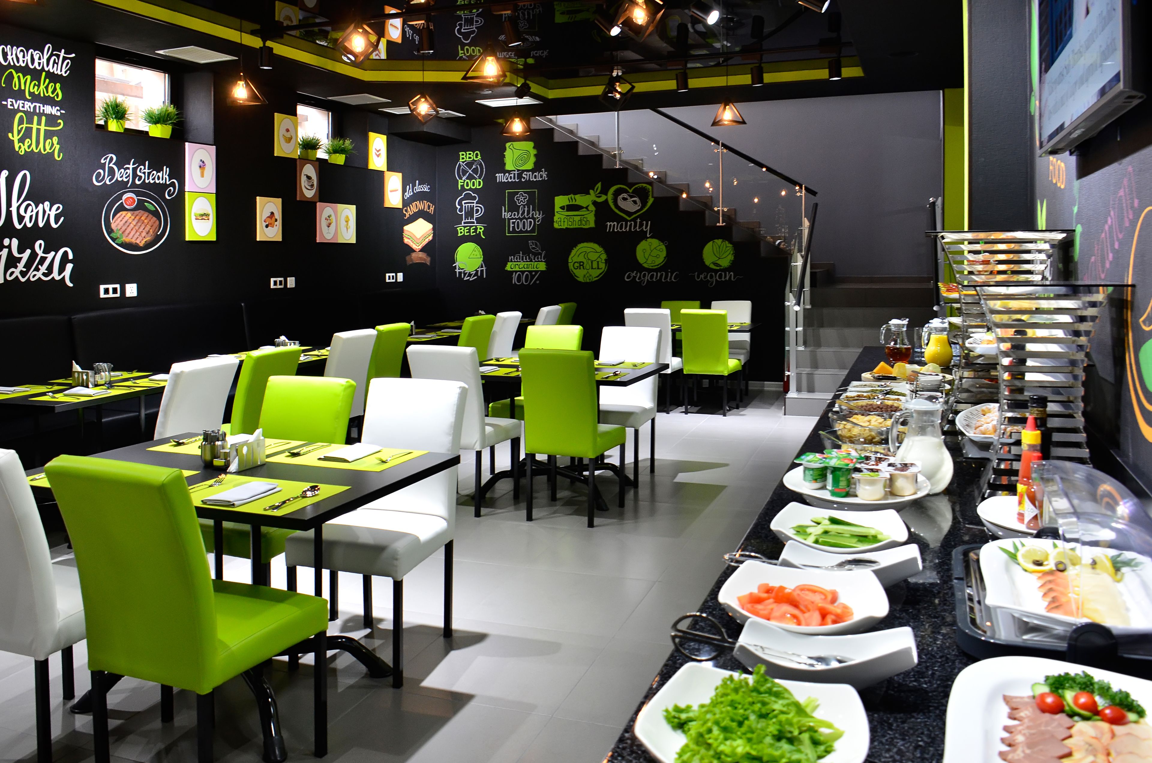 A modern restaurant with green and white chairs, a buffet with various food items, and walls decorated with food-related graphics and text.
