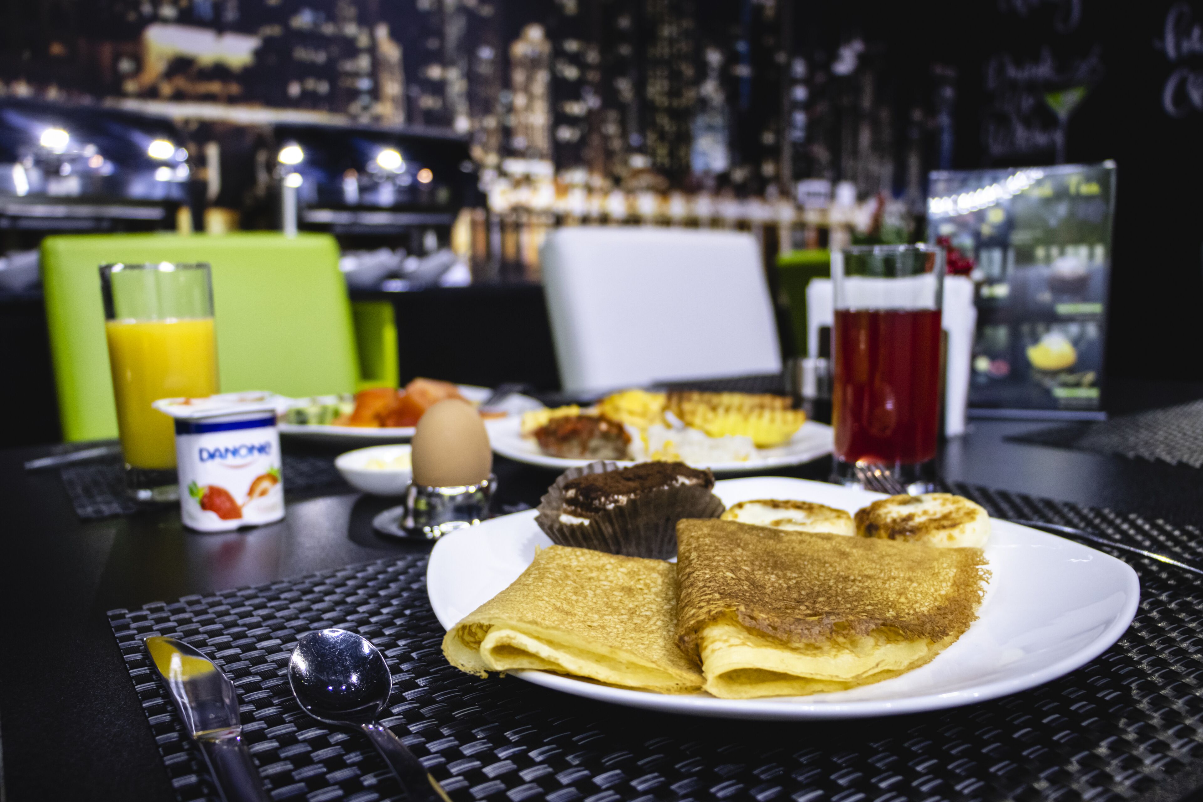 A breakfast setting with a plate of crepes, a glass of orange juice, and a cup of coffee on a table with a city view in the background.