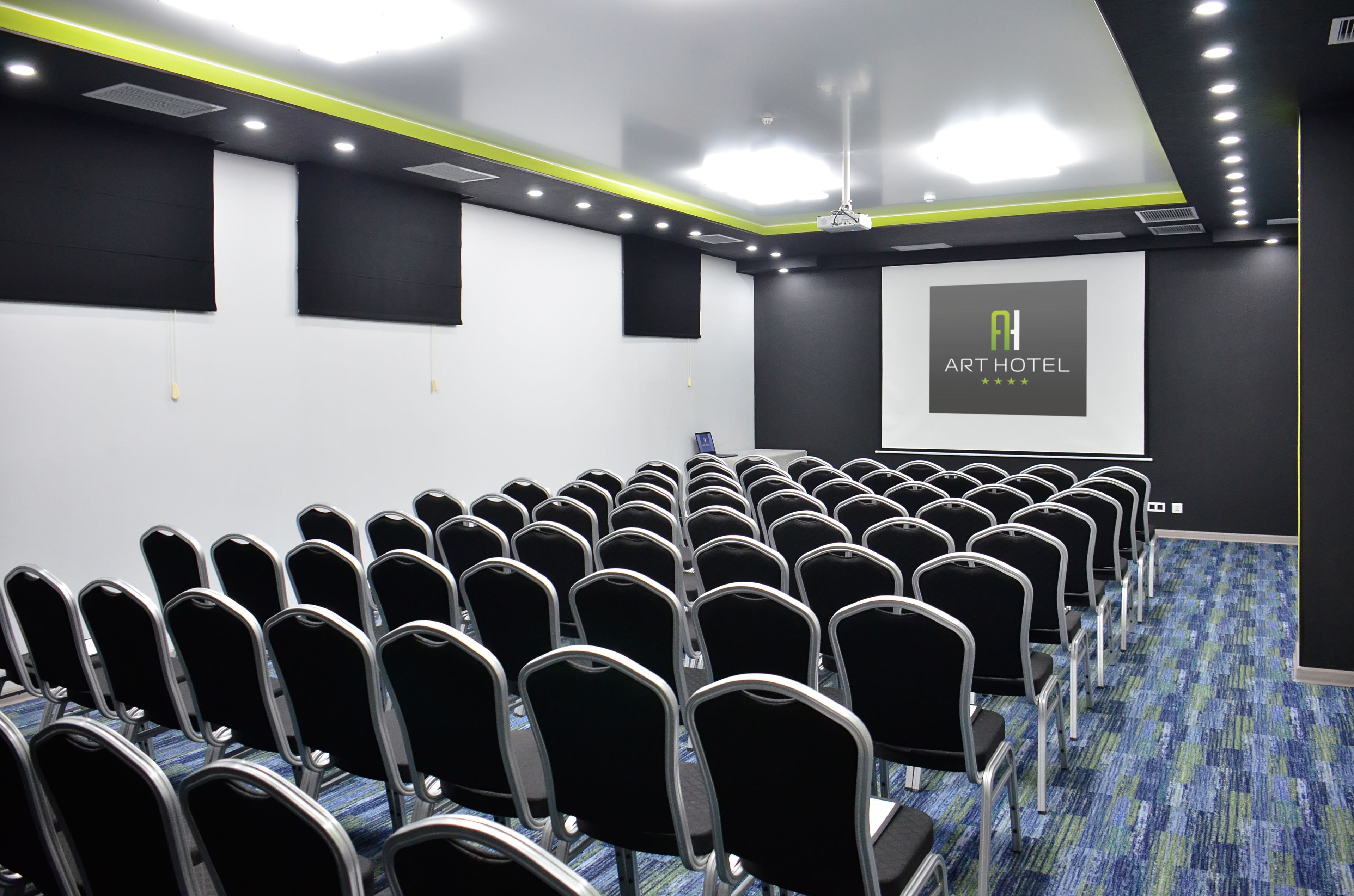Ein Konferenzraum mit Stuhlreihen, einer Projektionsleinwand, die "Art Hotel" zeigt, und einem Teppichboden.