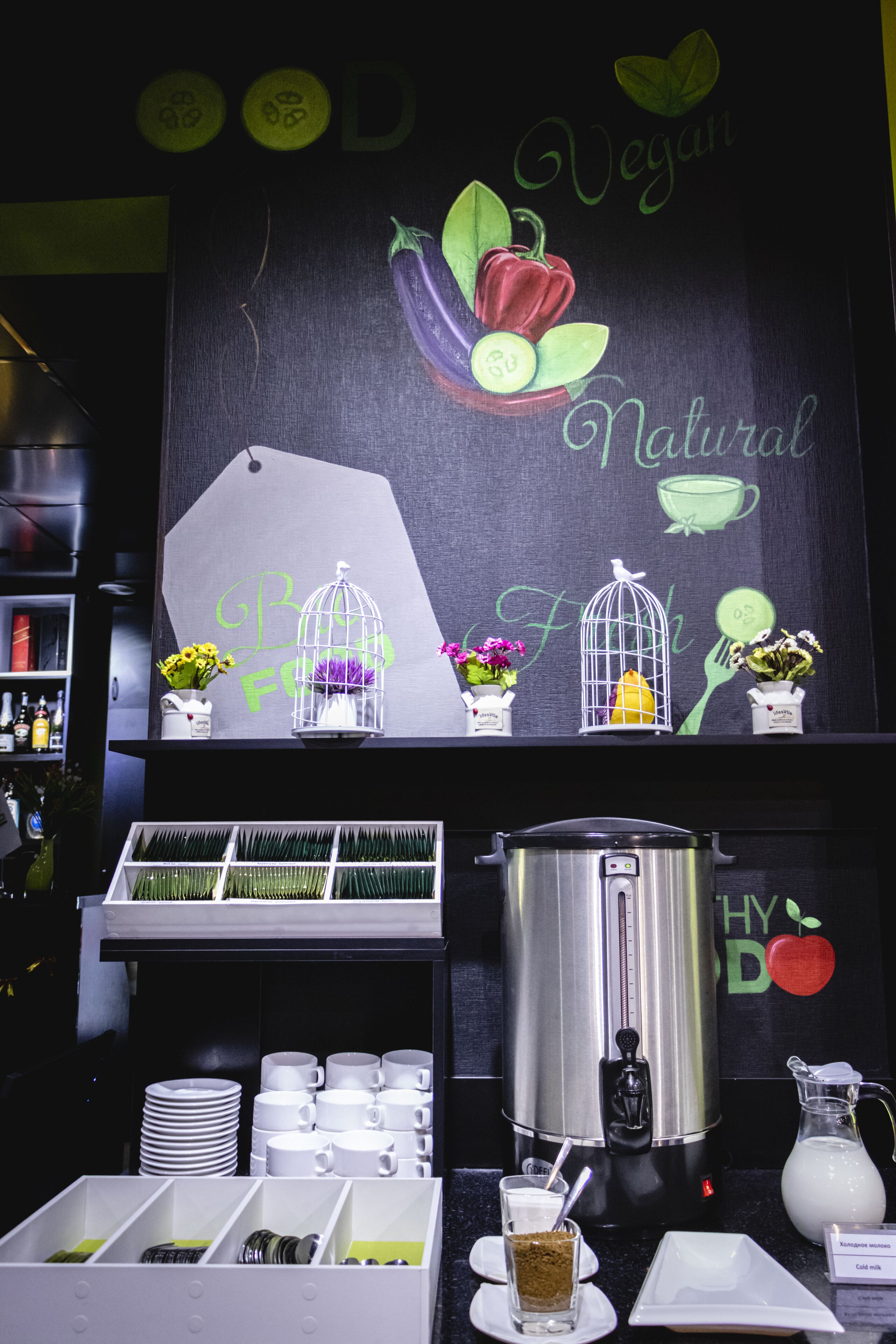 A breakfast bar with a chalkboard wall displaying vegan and natural food themes, a coffee machine, and a variety of breakfast items.