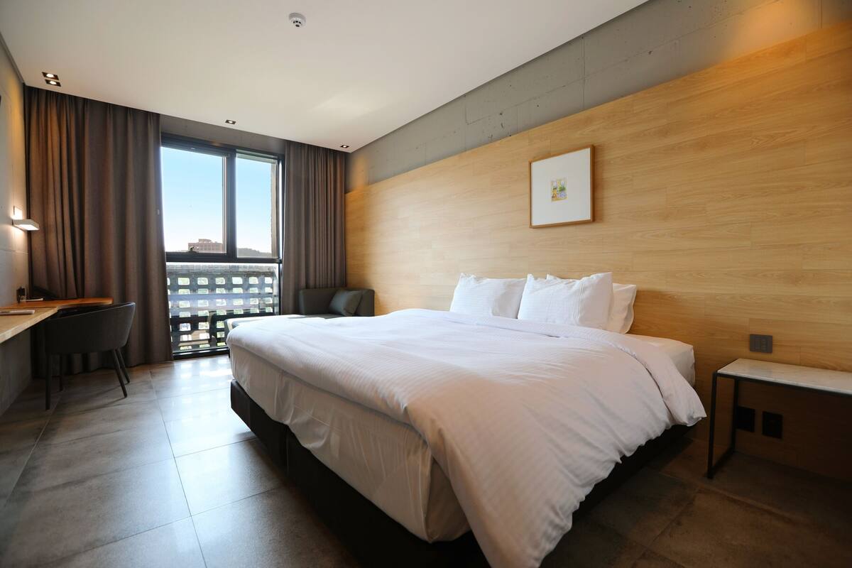 Une chambre d’hôtel moderne avec un grand lit, un bureau et une chaise. On y trouve une fenêtre avec des rideaux, un mur en bois et un tableau encadré.