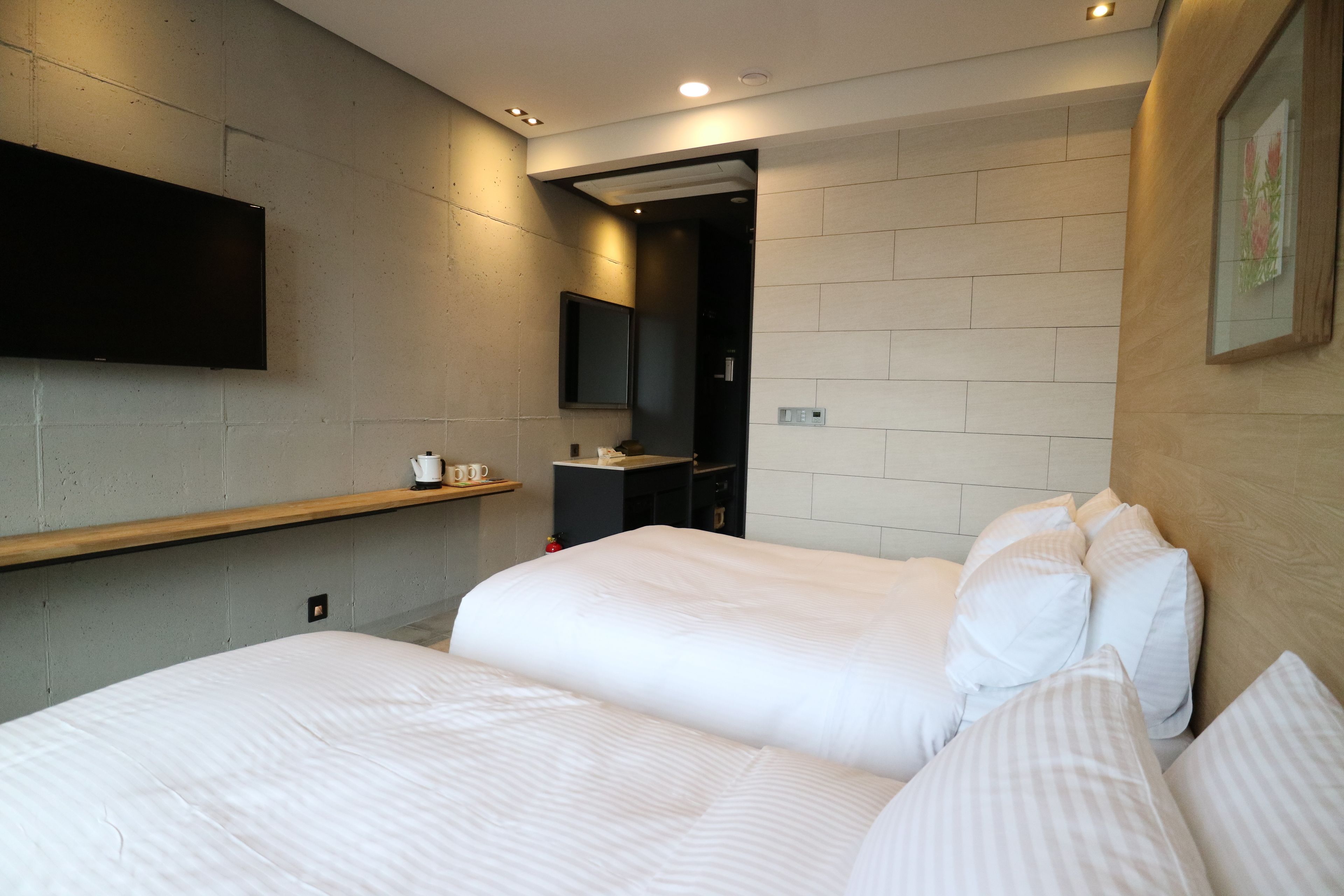 A modern hotel room with two beds, a mounted TV, a small shelf with a kettle and cups, and a wooden headboard.