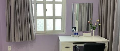 A room with a white desk, a black office chair, a window with grey curtains, a mirror, and a vase with flowers.