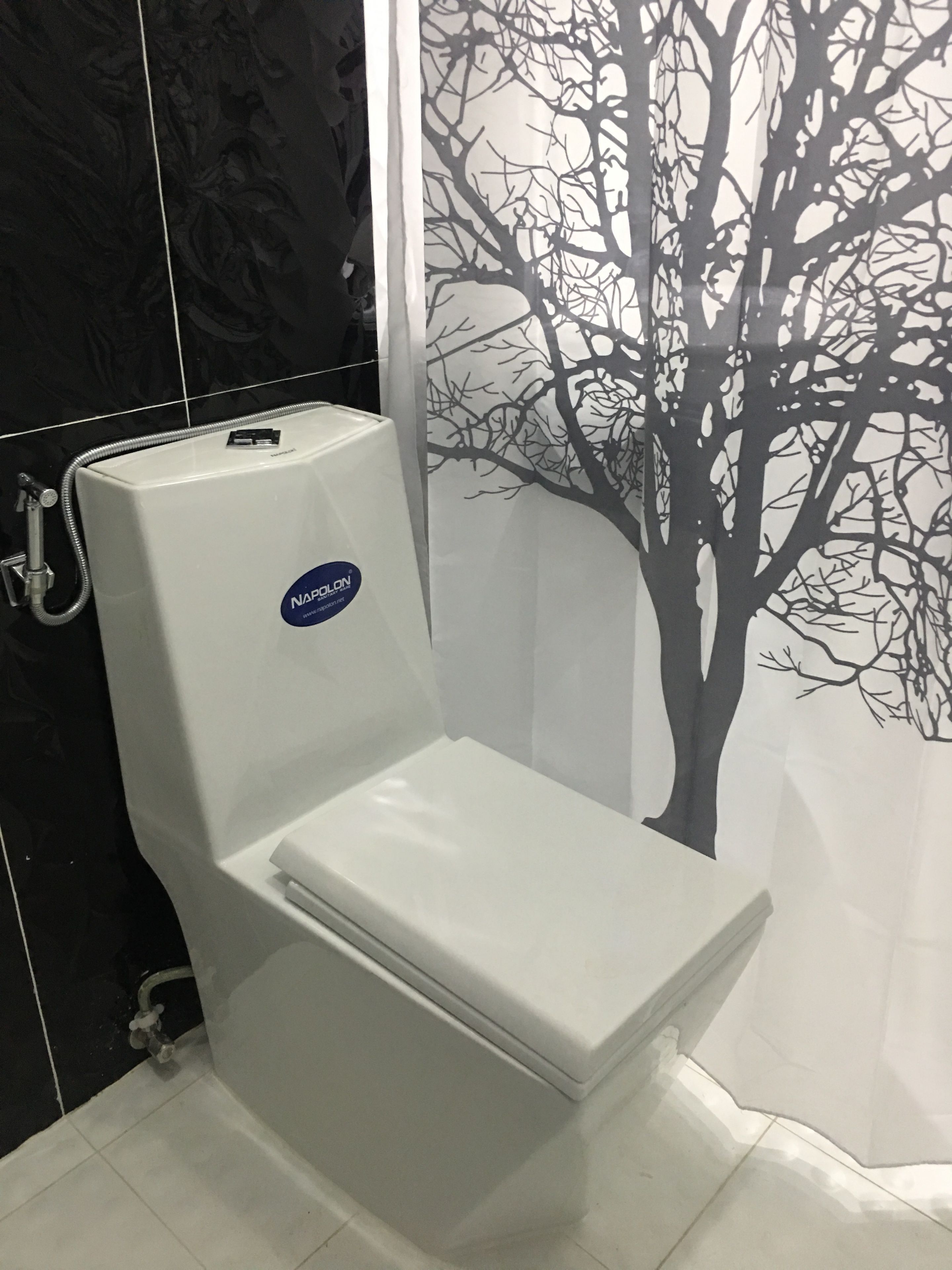 Des toilettes blanches avec une étiquette « NAPALON », un rideau de douche orné de motifs d’arbres et des murs recouverts de carreaux noirs.
