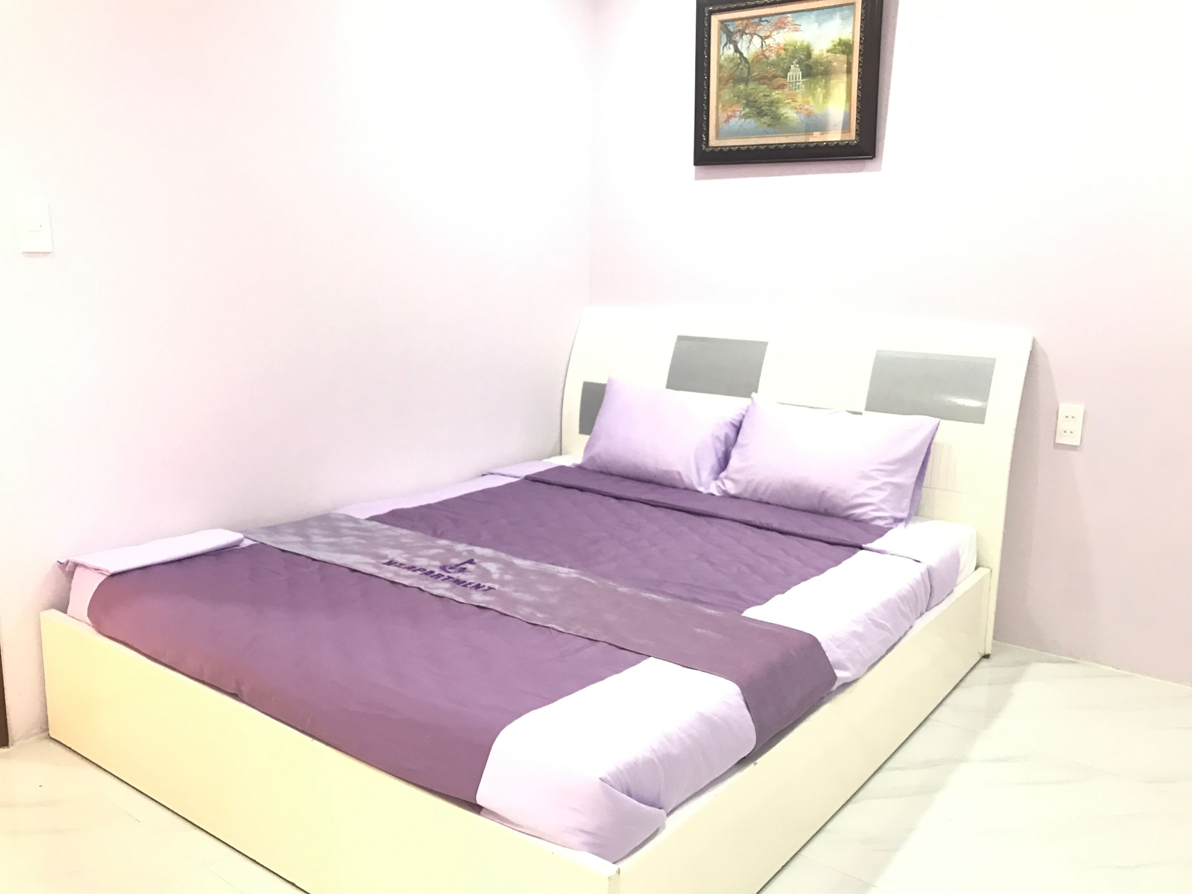 A bed with a purple bedspread and pillows, a white headboard, and a framed picture on the wall.