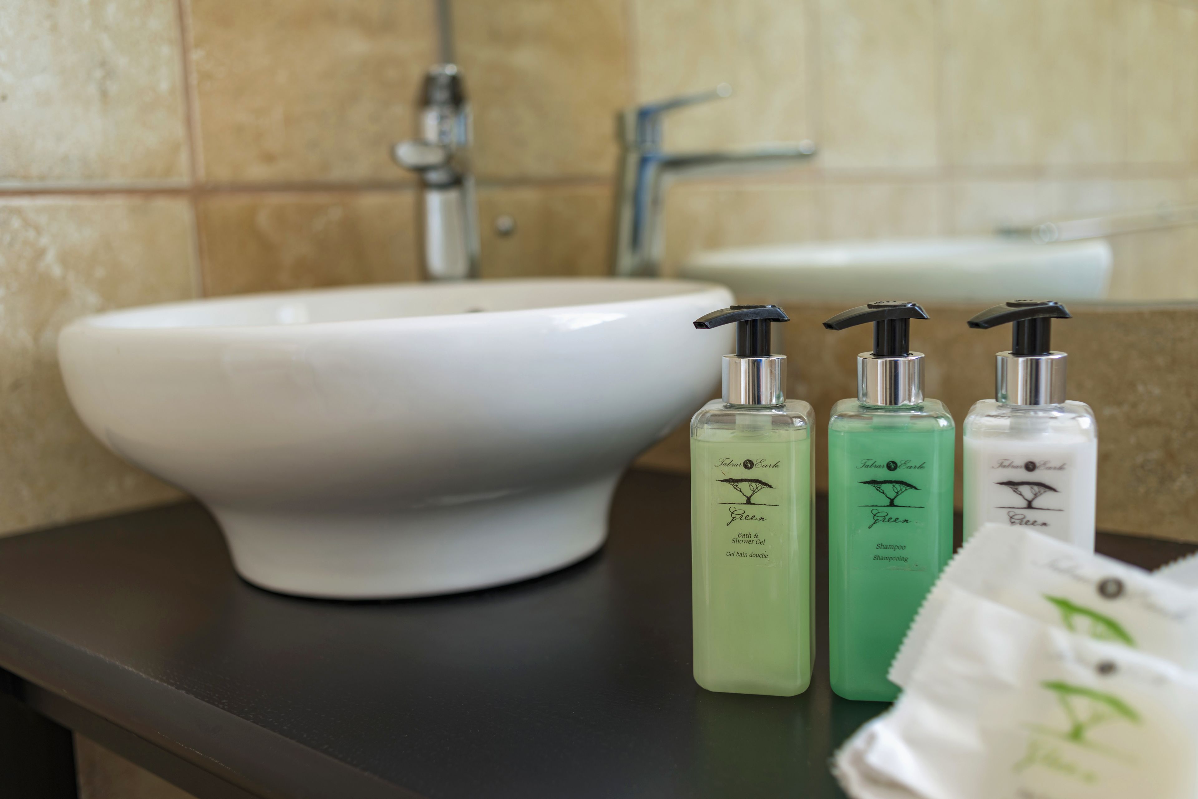A bathroom sink with three bottles of green liquid soap and a white towel on a dark countertop.