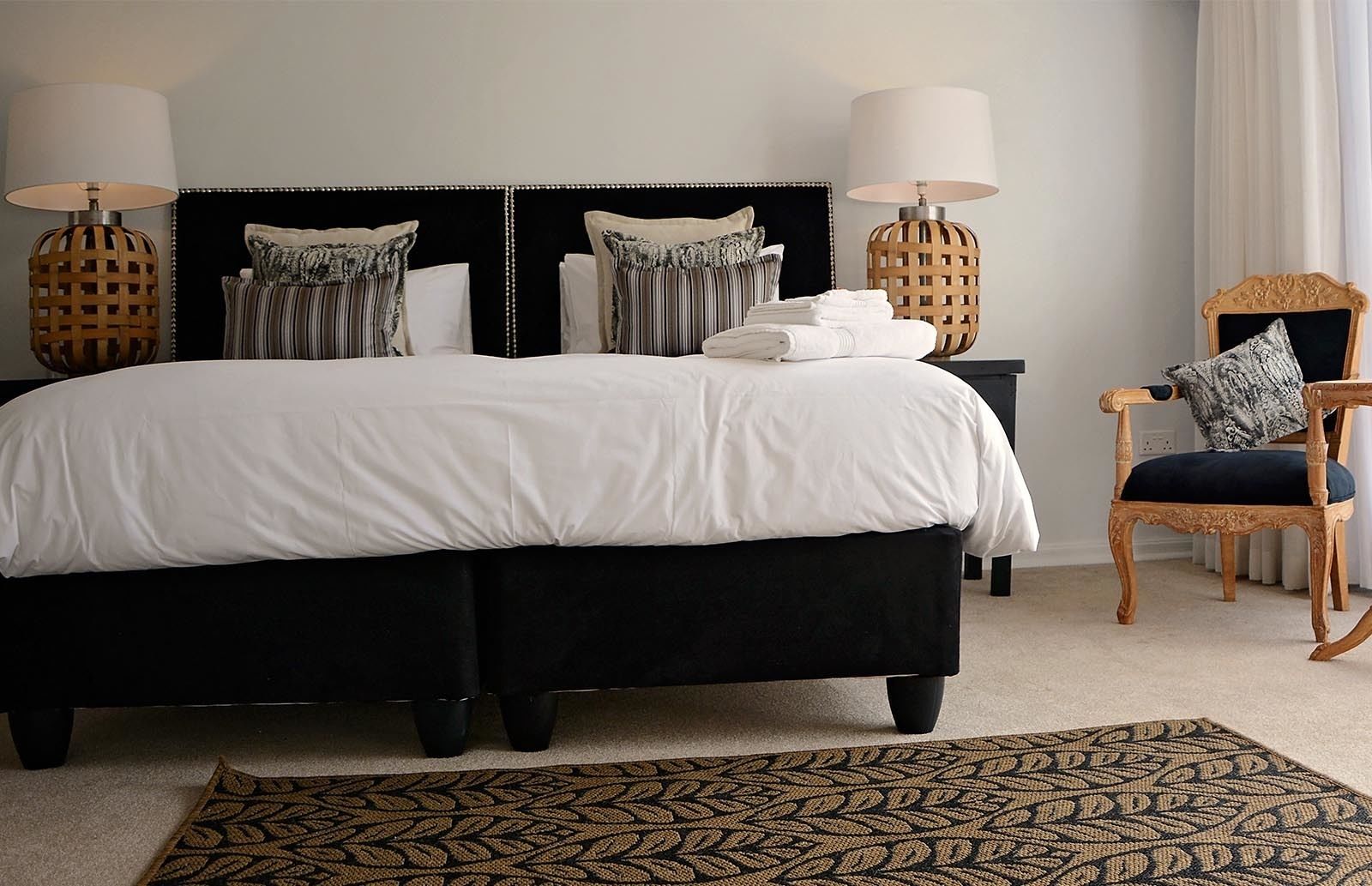 Un dormitorio con una cama tapizada de negro, dos mesitas de noche con lámparas, una silla de madera y una alfombra estampada.