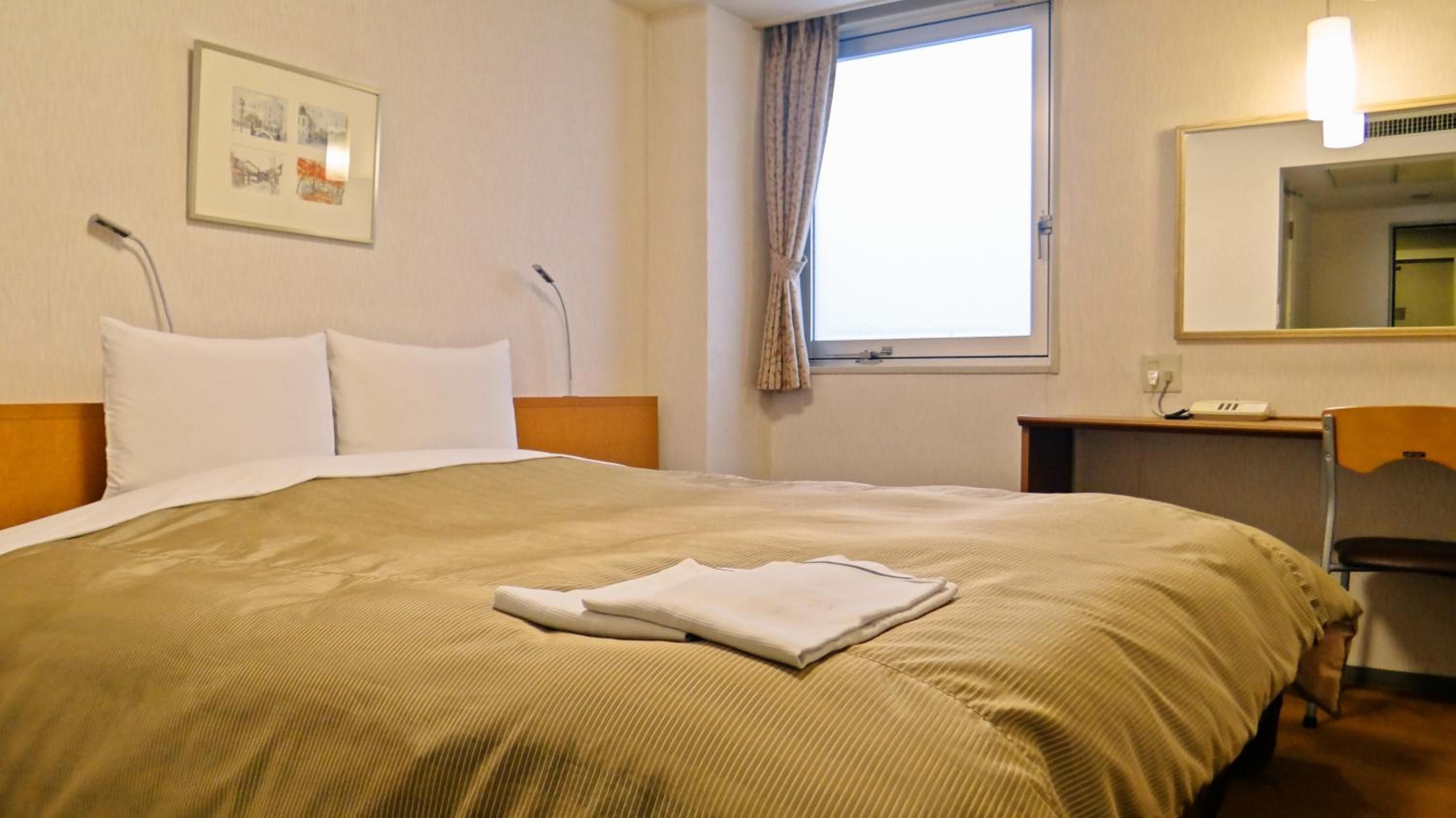 Une chambre d’hôtel comprenant un lit, une table de chevet, un bureau, une chaise, une fenêtre avec des rideaux, une lampe fixée au mur et un tableau encadré.