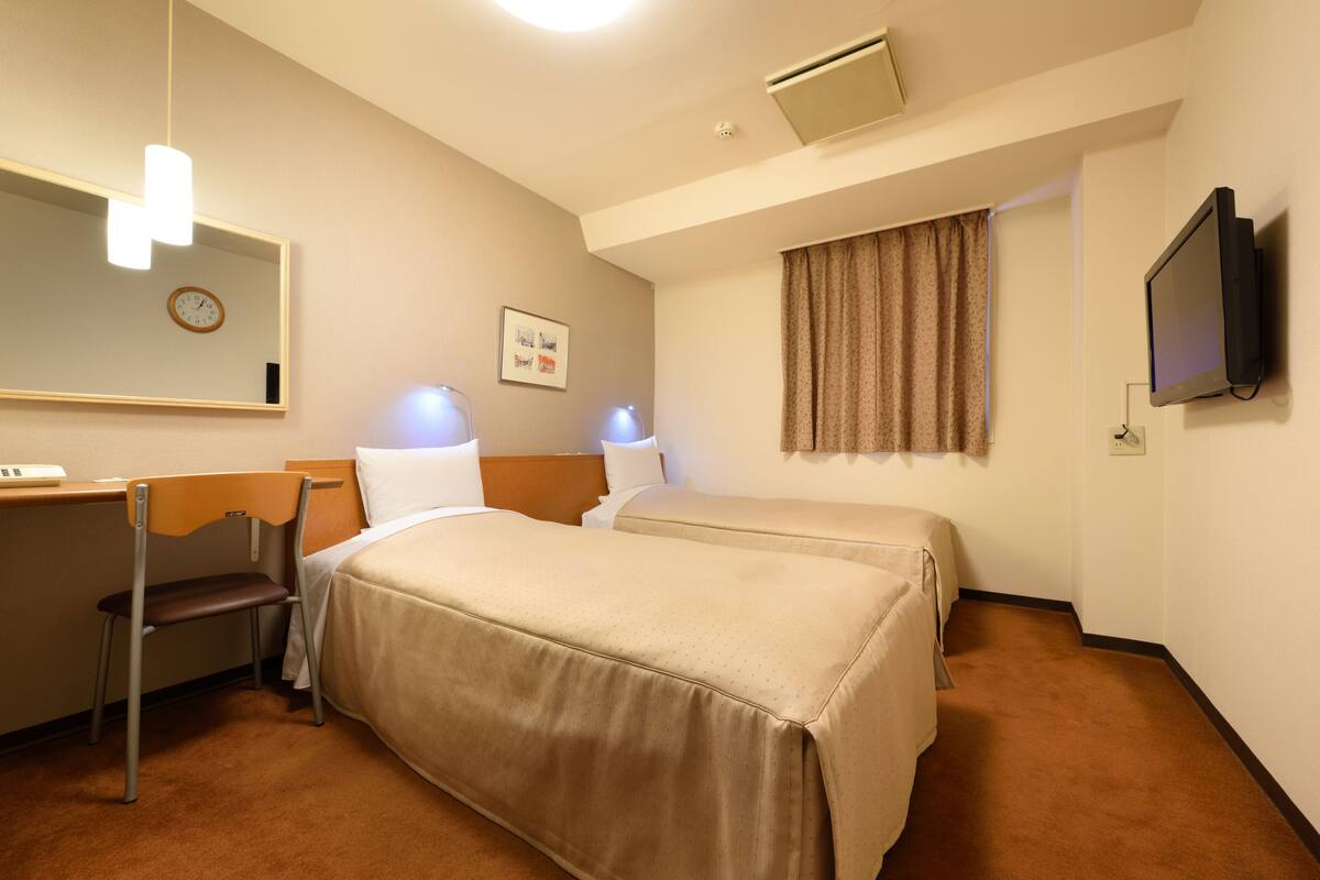 A hotel room with two beds, a desk, a chair, a clock, a television, and a window with curtains.