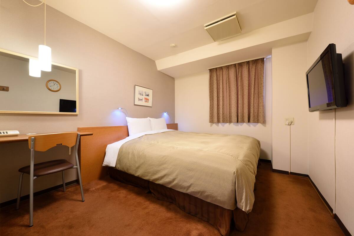 A hotel room with a bed, a desk with a chair, a television, a clock, and a window with curtains.