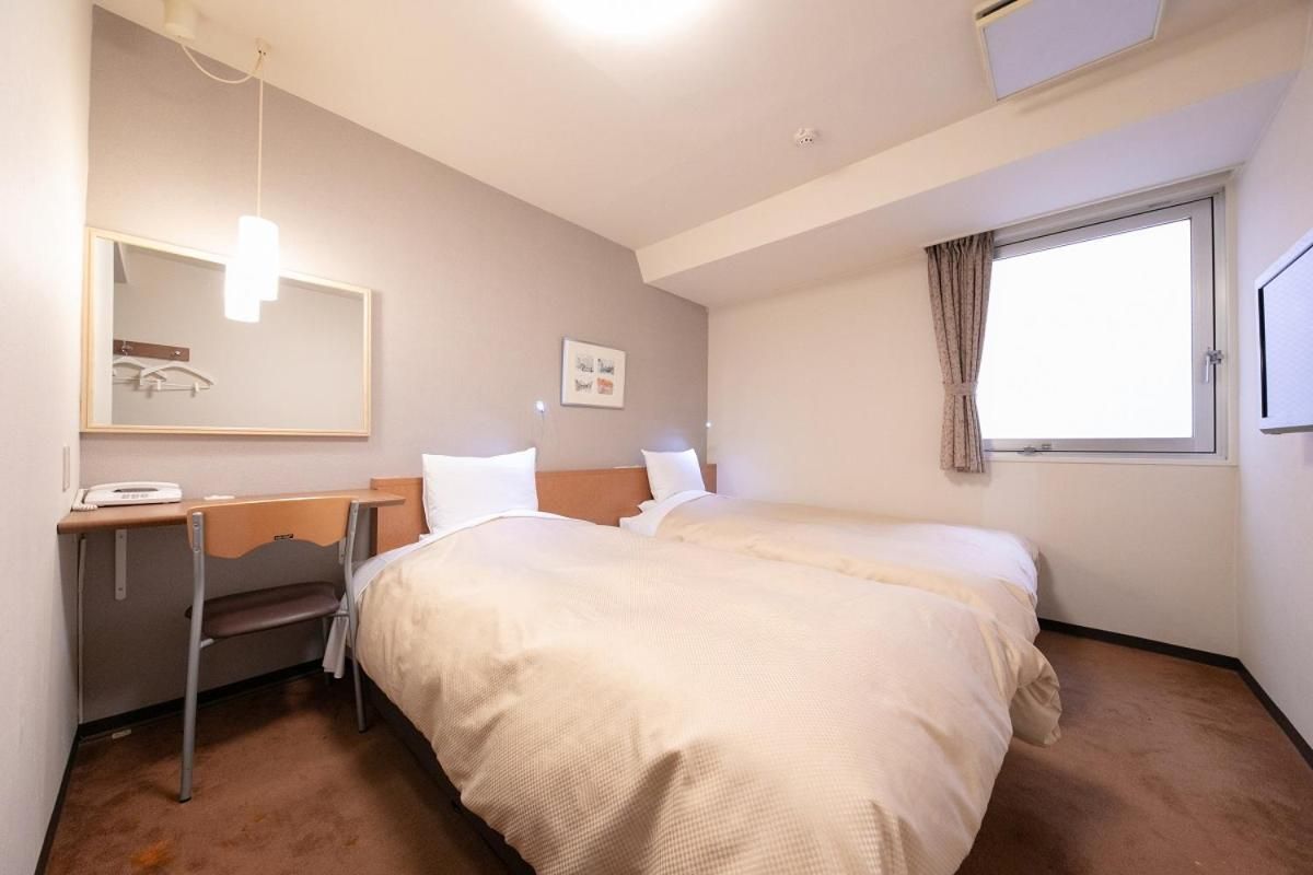 A hotel room with a bed, a desk, a chair, a mirror, a window, and a wall-mounted light fixture.