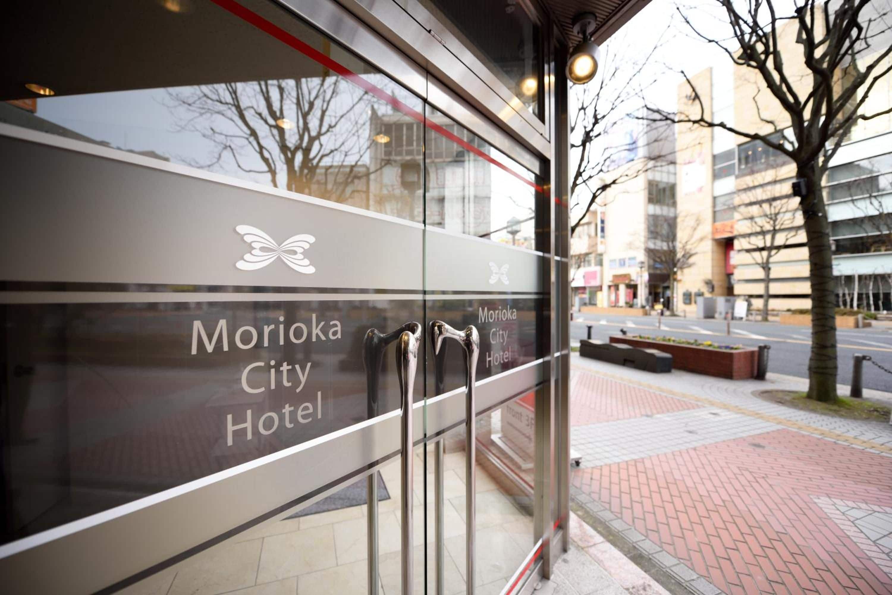 L'esterno dell'Hotel Morioka City con facciata in vetro, logo a forma di farfalla e vista chiara sulla strada e sugli edifici sullo sfondo.
