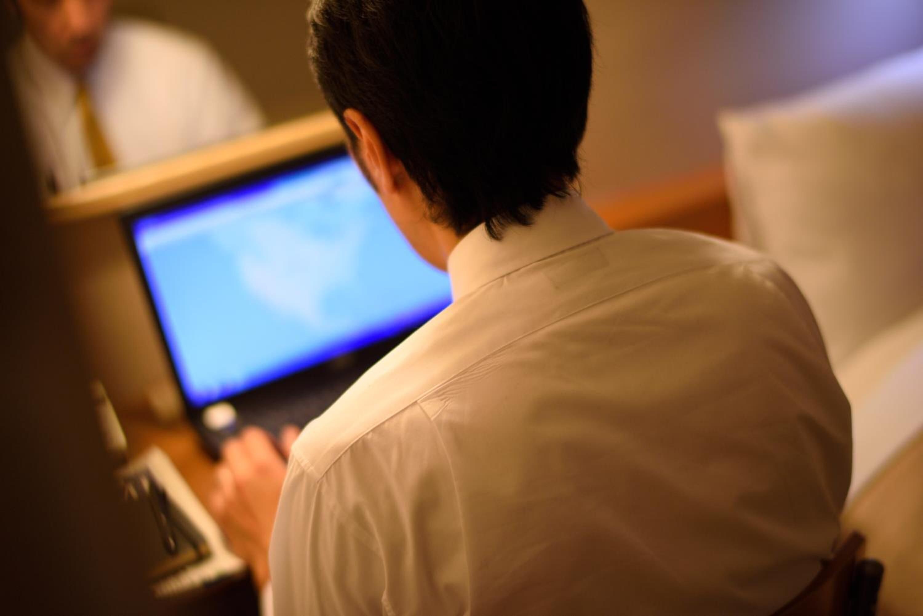 Una persona sentada en un escritorio con una laptop, en una habitación con una silla y una mesa.