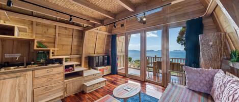 A wooden cabin interior with a kitchen, dining area, and a large sliding glass door leading to a balcony with a view of the ocean.
