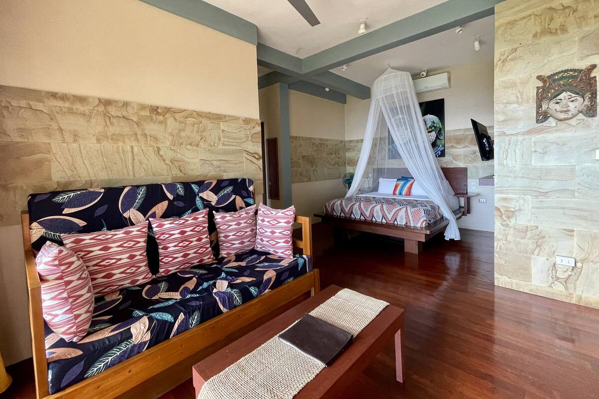 Een kamer met een bank en een bed, beide met beddengoed met een patroon. Er is een plafondventilator, een wandairconditioner en een decoratief kunstwerk aan de muur.