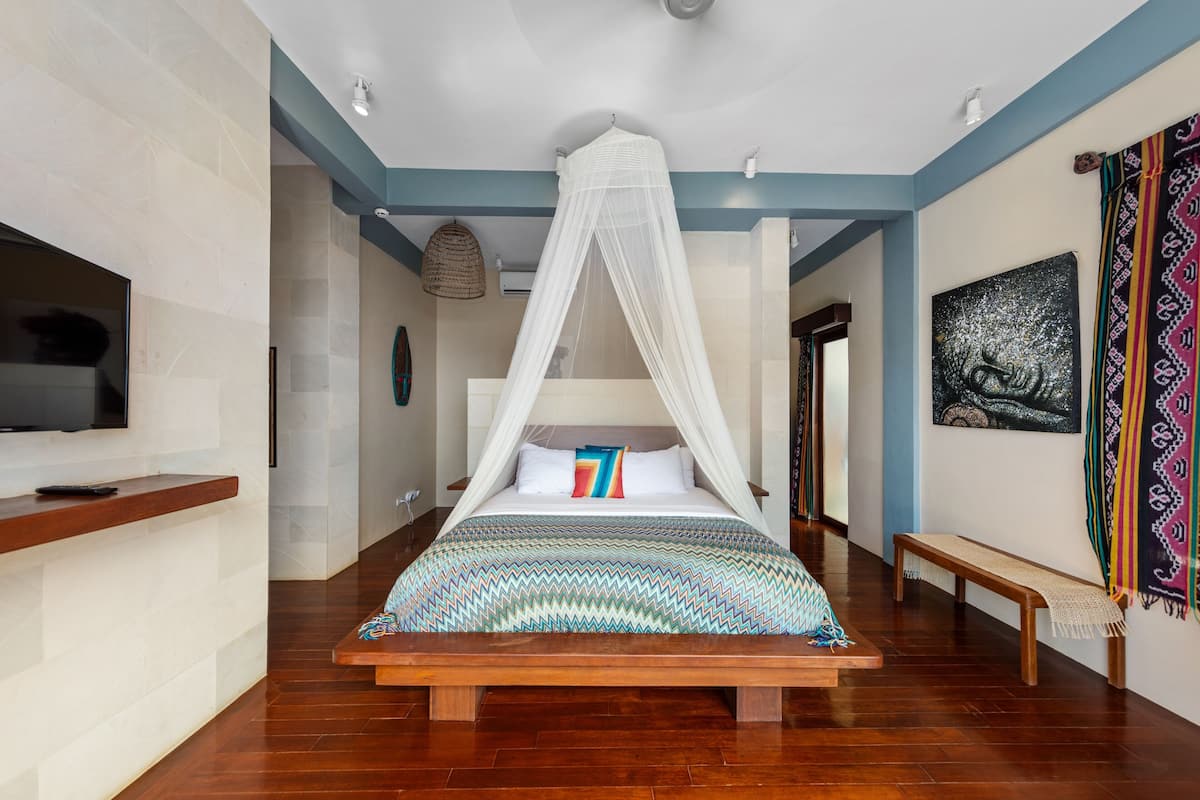 Een slaapkamer met een bed onder een witte hemel, een houten bankje, een televisie op een standaard en een wand met een wandtapijt.