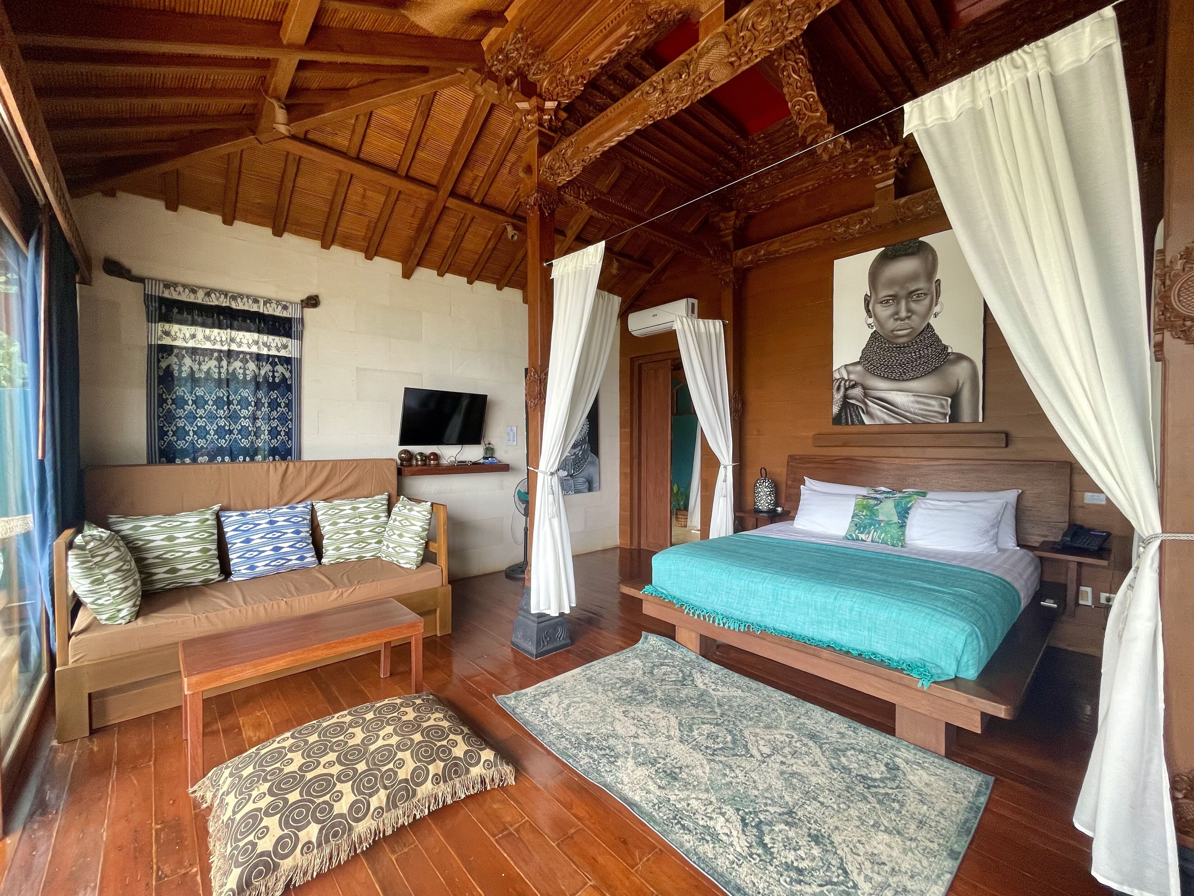 A room with a wooden ceiling, a bed with a green bedspread, a sofa with patterned cushions, a bench, and a patterned rug.