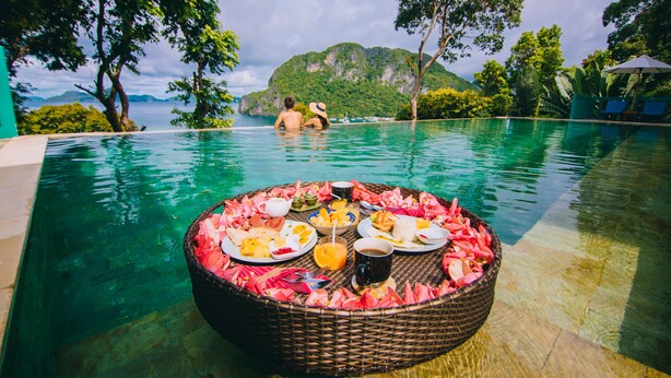 A poolside dining setup with a wicker table surrounded by plates of food and drinks, overlooking a tropical landscape with mountains and a clear sky.