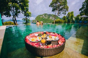 Une table en osier installée au bord de la piscine, entourée d’assiettes remplies de plats et de boissons, d’où l’on peut admirer un paysage tropical avec des montagnes et un ciel dégagé.
