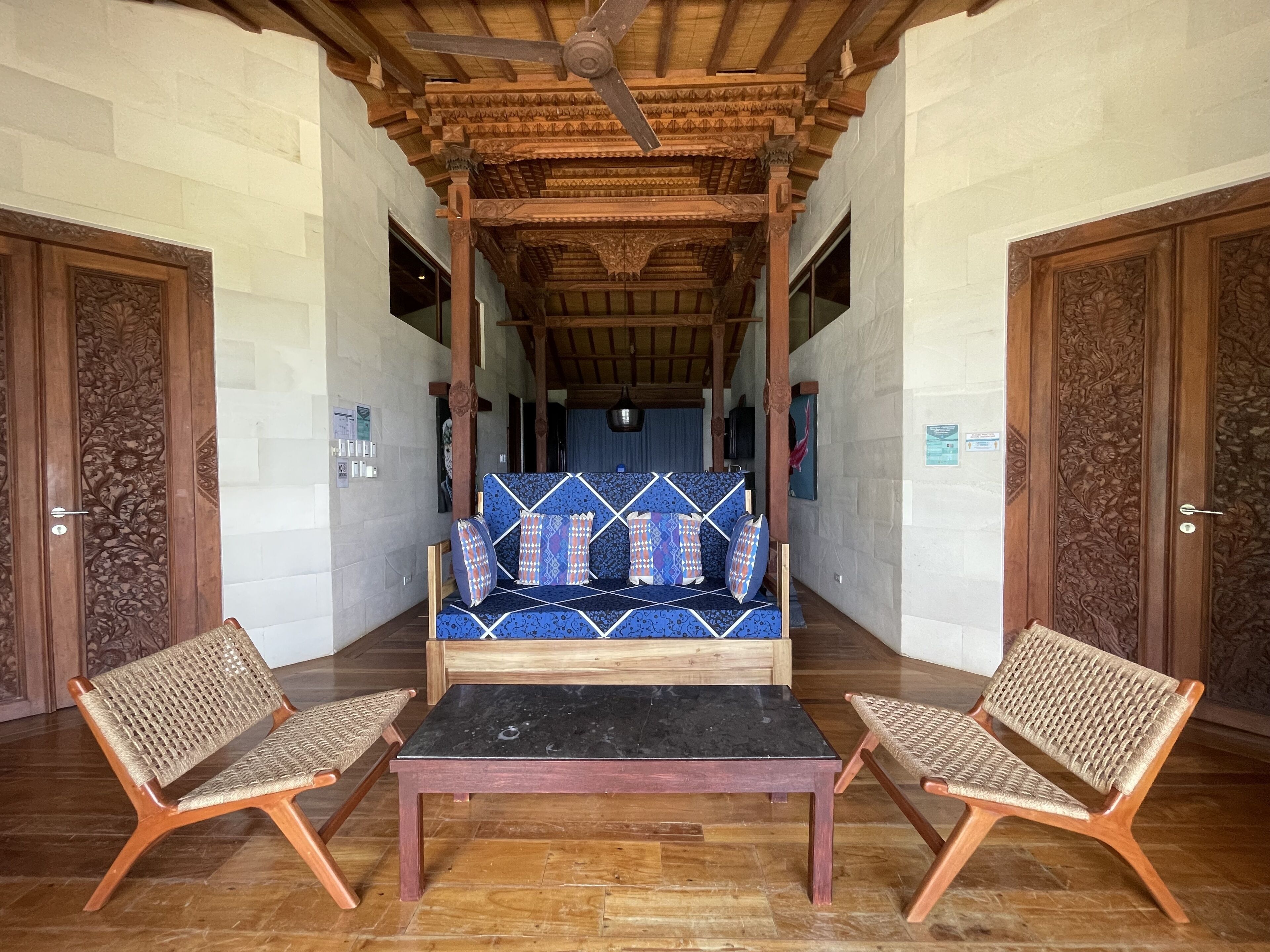 Una habitación con un sofá central, dos sillones de mimbre, una mesa negra y puertas de madera con intrincados grabados.