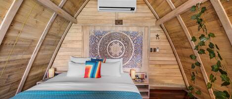 A bedroom with a sloped ceiling, a bed with blue bedding, a decorative wall tapestry, and an air conditioning unit.