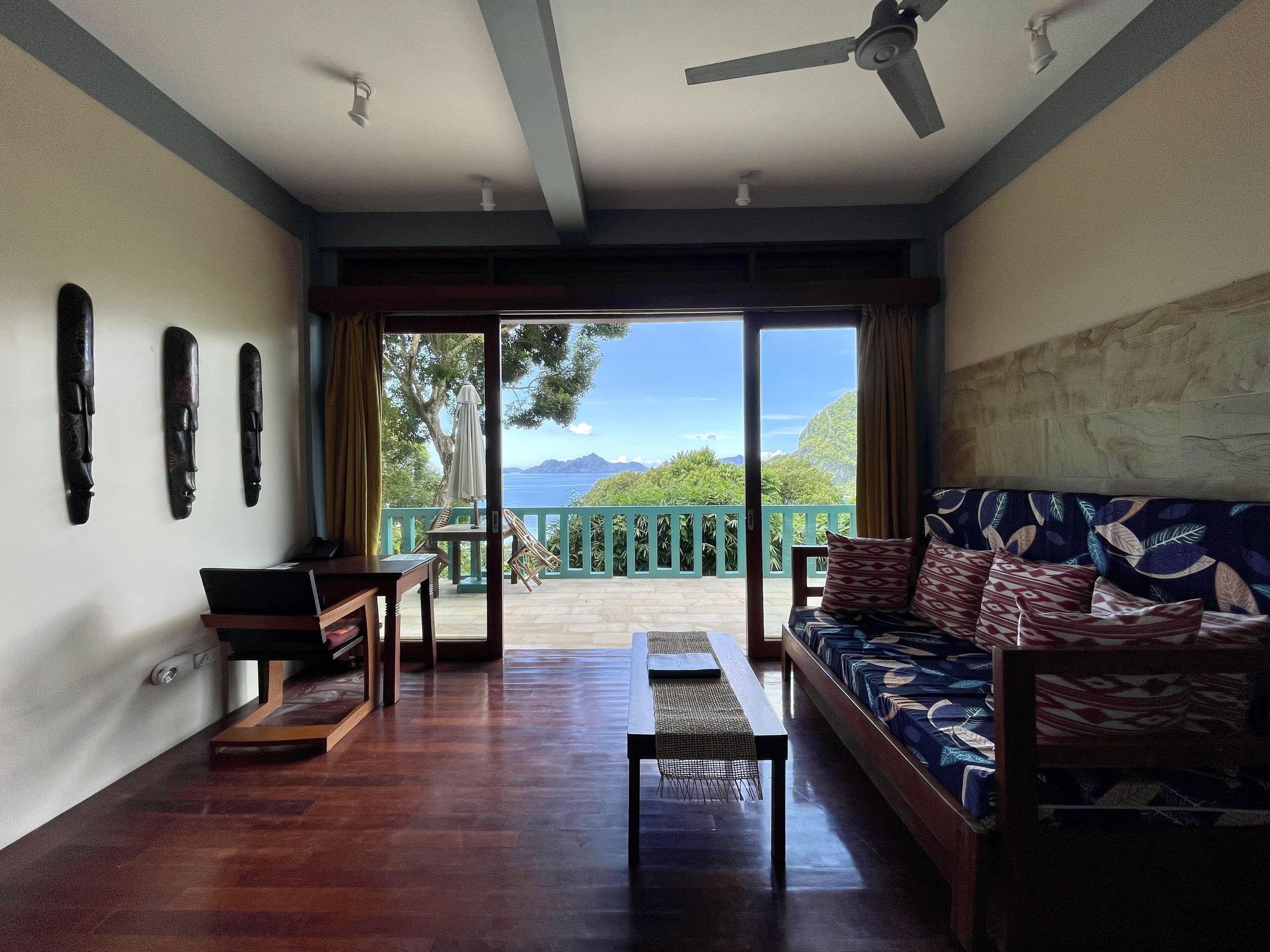 A spacious room with a wooden floor, a sofa with patterned cushions, a table, and a laptop. A ceiling fan is visible, and there are three wall-mounted items. The room has a large sliding glass door leading to a balcony with a view of the sea and mountains.