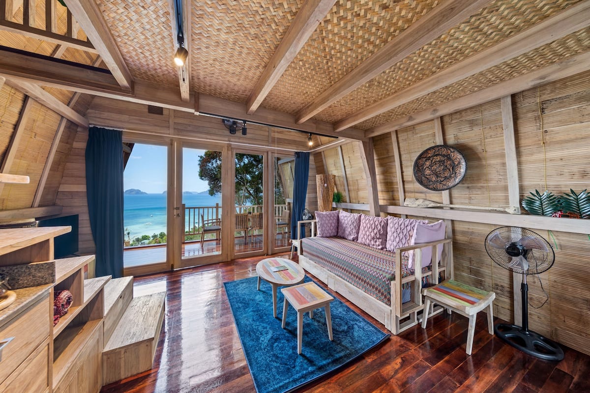 Een gezellig, houten interieur met een groot raam met uitzicht op zee, een bank, een kleine tafel en een ventilator.