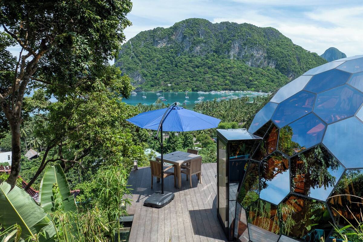 Een modern terras buiten met een blauwe parasol, omgeven door weelderige begroeiing en uitzicht op een baai met boten.