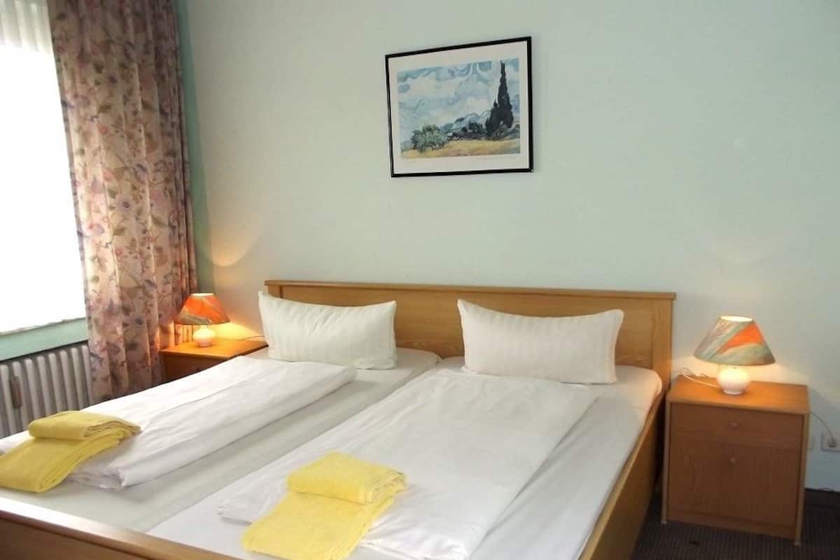 A double bed with white linens and two pillows, a wooden headboard, a nightstand with a lamp, and a framed painting on the wall.