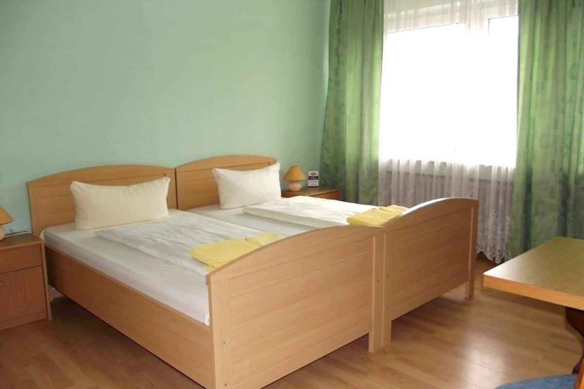 A bedroom with a wooden bed, two pillows, a bedside table with a lamp, and a window with green curtains.