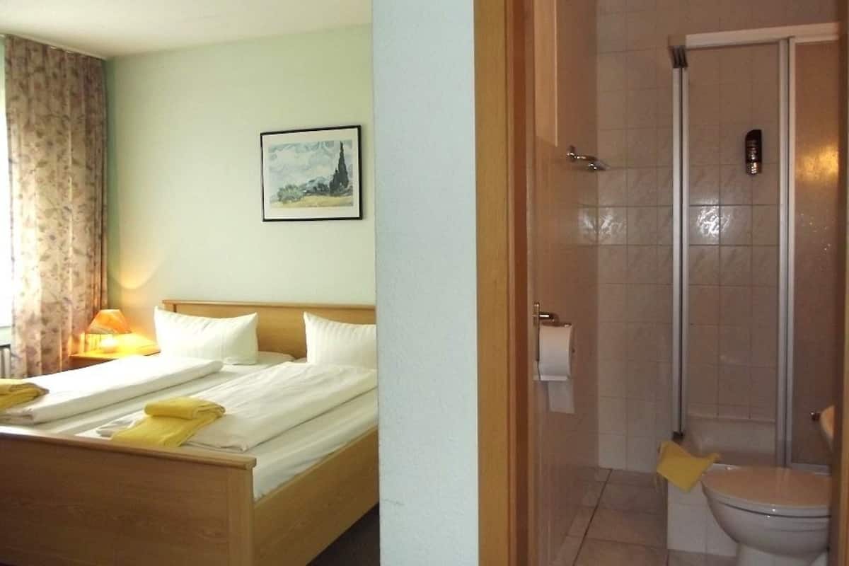 A hotel room with a bed, a bedside table with a lamp, a bathroom with a shower, and a toilet.