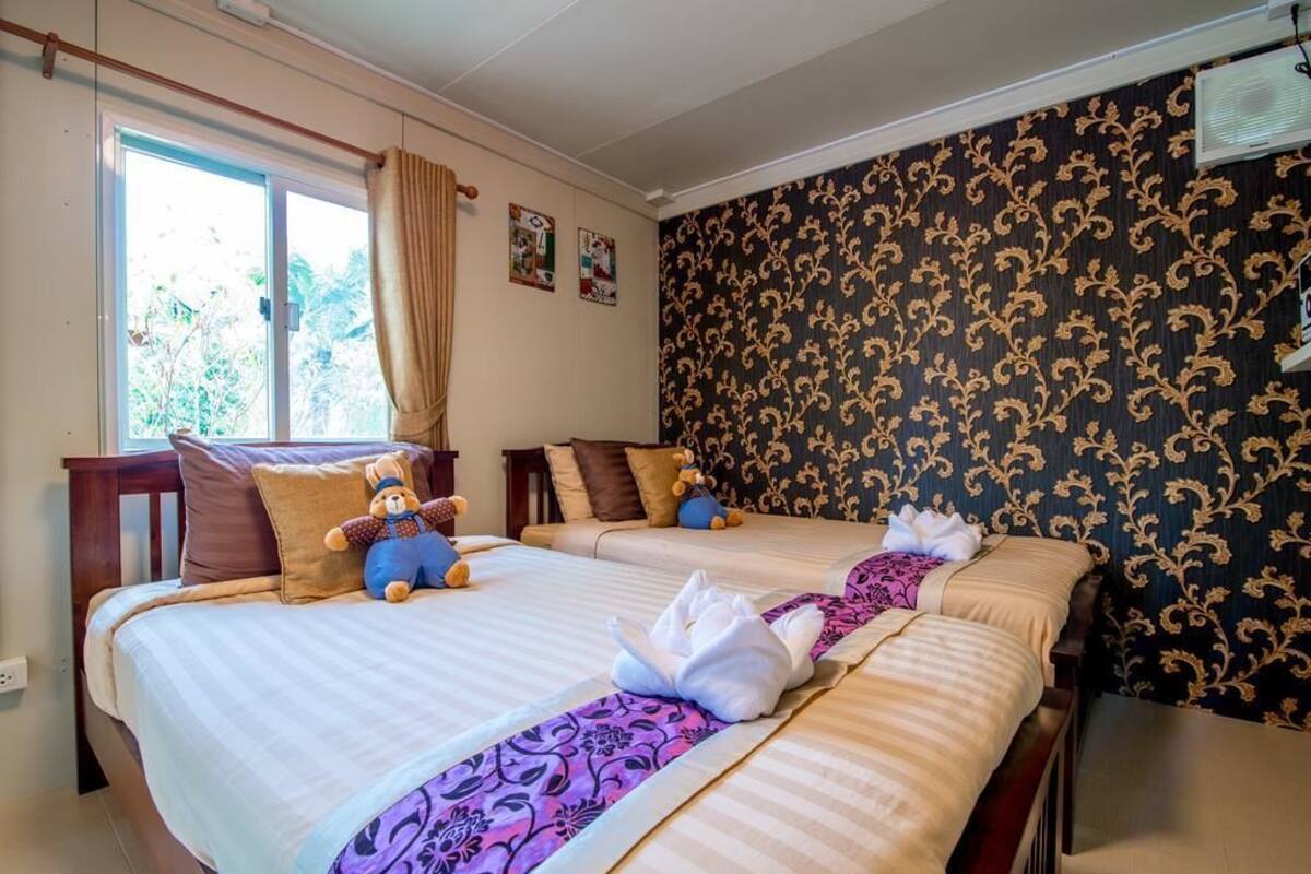 Ein Hotelzimmer mit zwei Betten, einem Fenster mit Vorhängen, einer Wand mit ornamentalem Wandpapier und einem kleinen Tisch mit einer Lampe.